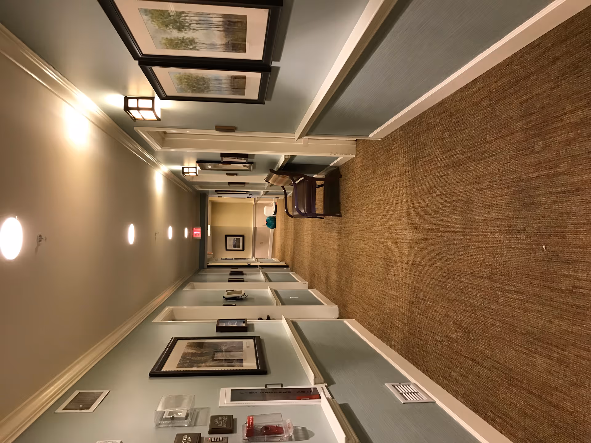 A well-lit hallway in a senior living facility with light blue walls, brown carpet, framed artwork on the walls, and a wooden bench on the right side. There are several doors along the hallway and ceiling lights evenly spaced. An exit sign is visible at the far end of the hallway.
