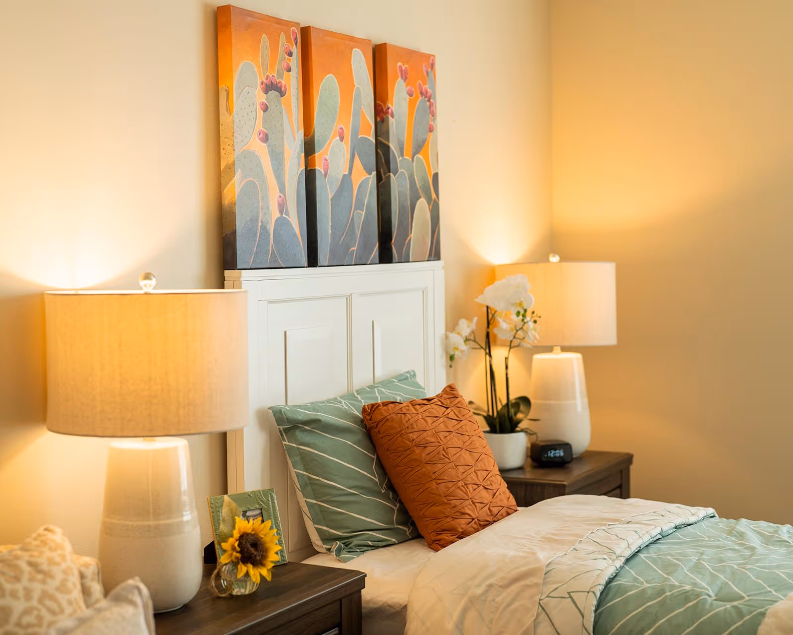 A cozy bedroom with a white headboard, decorative pillows and bedding, two bedside tables with lamps and a three-panel cactus artwork above the bed.