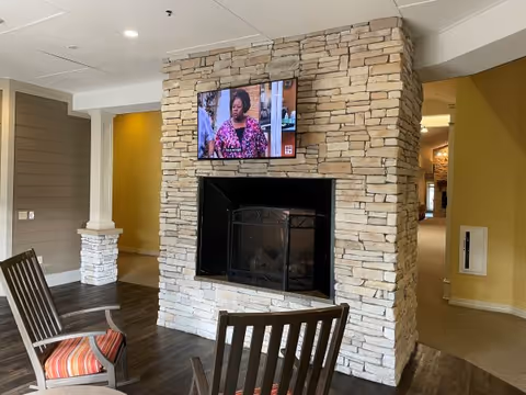 A cozy living area featuring a stone fireplace with a black metal screen. Above the fireplace, a flat-screen TV is mounted showing a woman in a pink patterned top. Two wooden chairs with striped cushions are positioned facing the fireplace. The room has wood flooring and walls painted in neutral tones with yellow accents. A hallway is visible to the right.