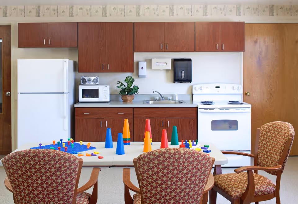 A kitchen area in a senior living facility with wooden cabinets, a white refrigerator, microwave, stove, and a sink. In front of the kitchen is a table with colorful plastic cones and pegs arranged on it, surrounded by four patterned chairs.
