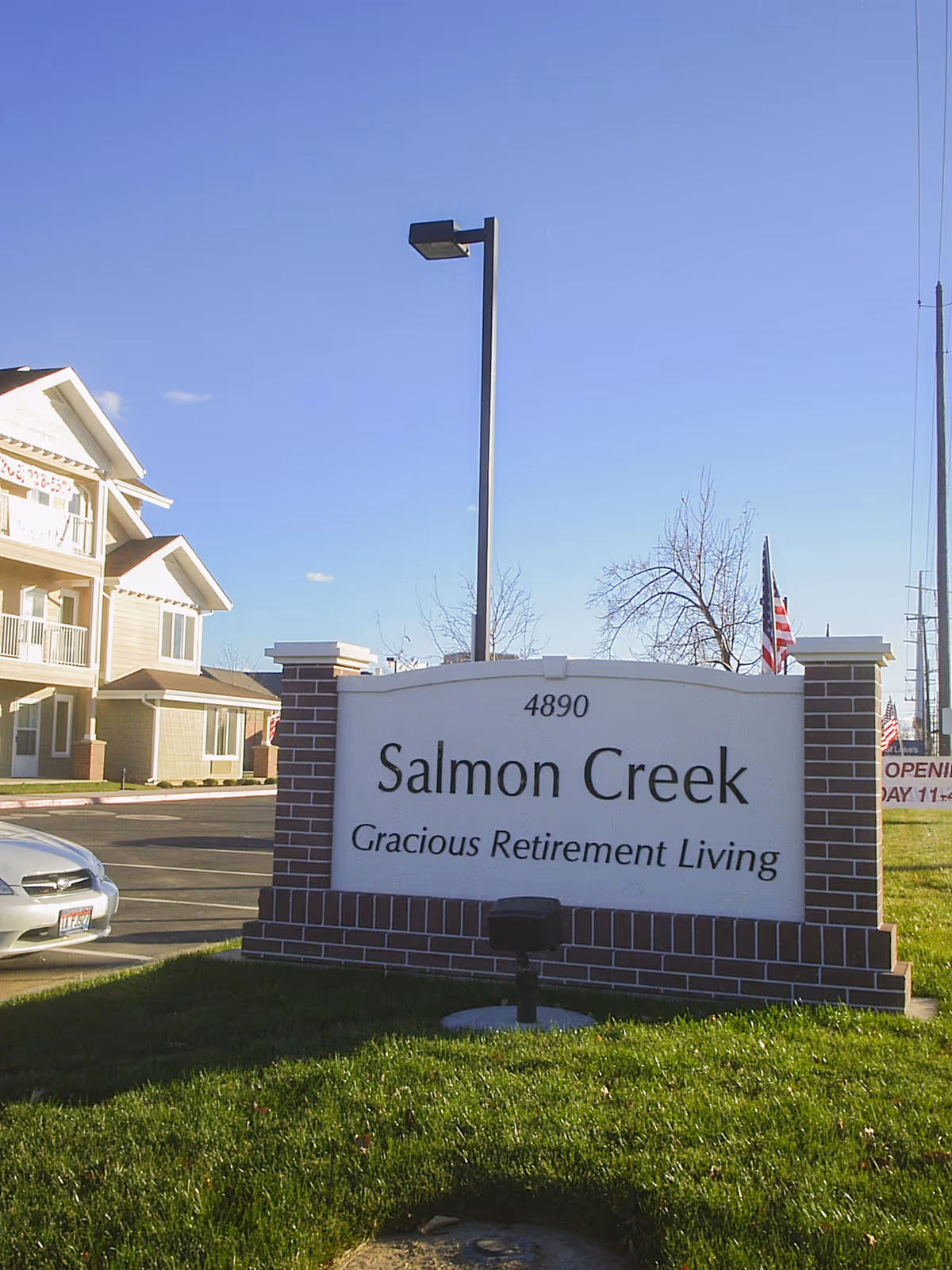 Outdoor view of a brick and white sign for Salmon Creek Gracious Retirement Living with a building, parked car, and clear blue sky in the background.