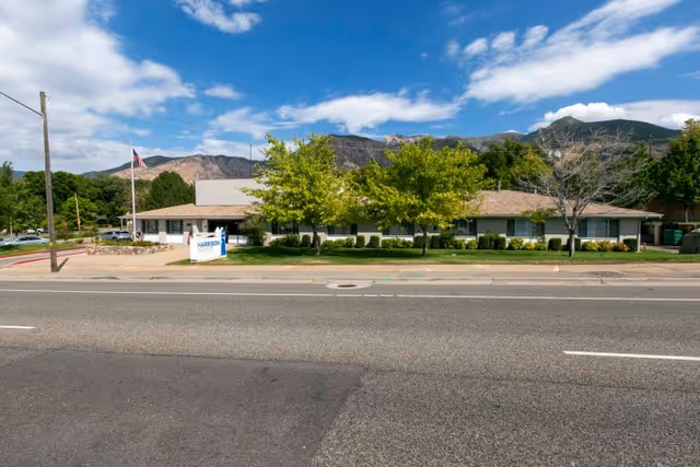 Exterior view of Harrison Pointe Healthcare And Rehabilitation building with a clear blue sky, some clouds, trees in front, and mountains in the background. There is a sign with the facility name near the entrance and an American flag on a pole.