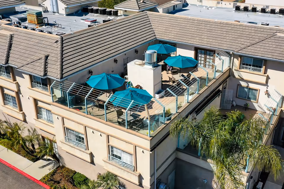 Aerial view of a rooftop patio area on a beige multi-story building with several teal umbrellas providing shade over tables and chairs. The patio is enclosed with glass railings and surrounded by a tiled roof. Palm trees and landscaping are visible around the building.