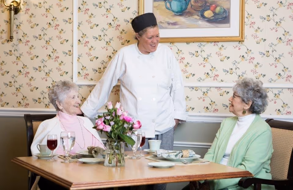 A chef in a white uniform and black hat stands smiling and talking to two elderly women seated at a dining table set with plates, glasses, and a vase of pink flowers in a room with floral wallpaper and a framed painting.