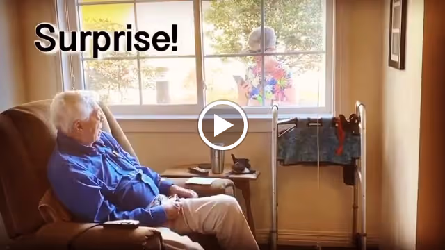 An elderly man sitting in a recliner chair inside a room, looking out a window where another person is standing outside holding a phone. The word 'Surprise!' is displayed on the window.