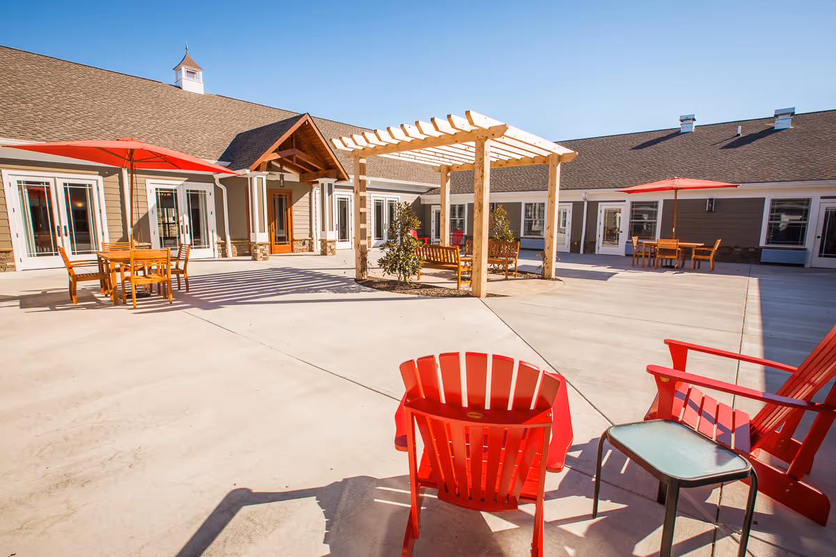 Outdoor patio area at Signature Lifestyles of Rogersville featuring red Adirondack chairs, wooden tables with red umbrellas, a wooden pergola with benches underneath, and a building with multiple doors and windows in the background under a clear blue sky.
