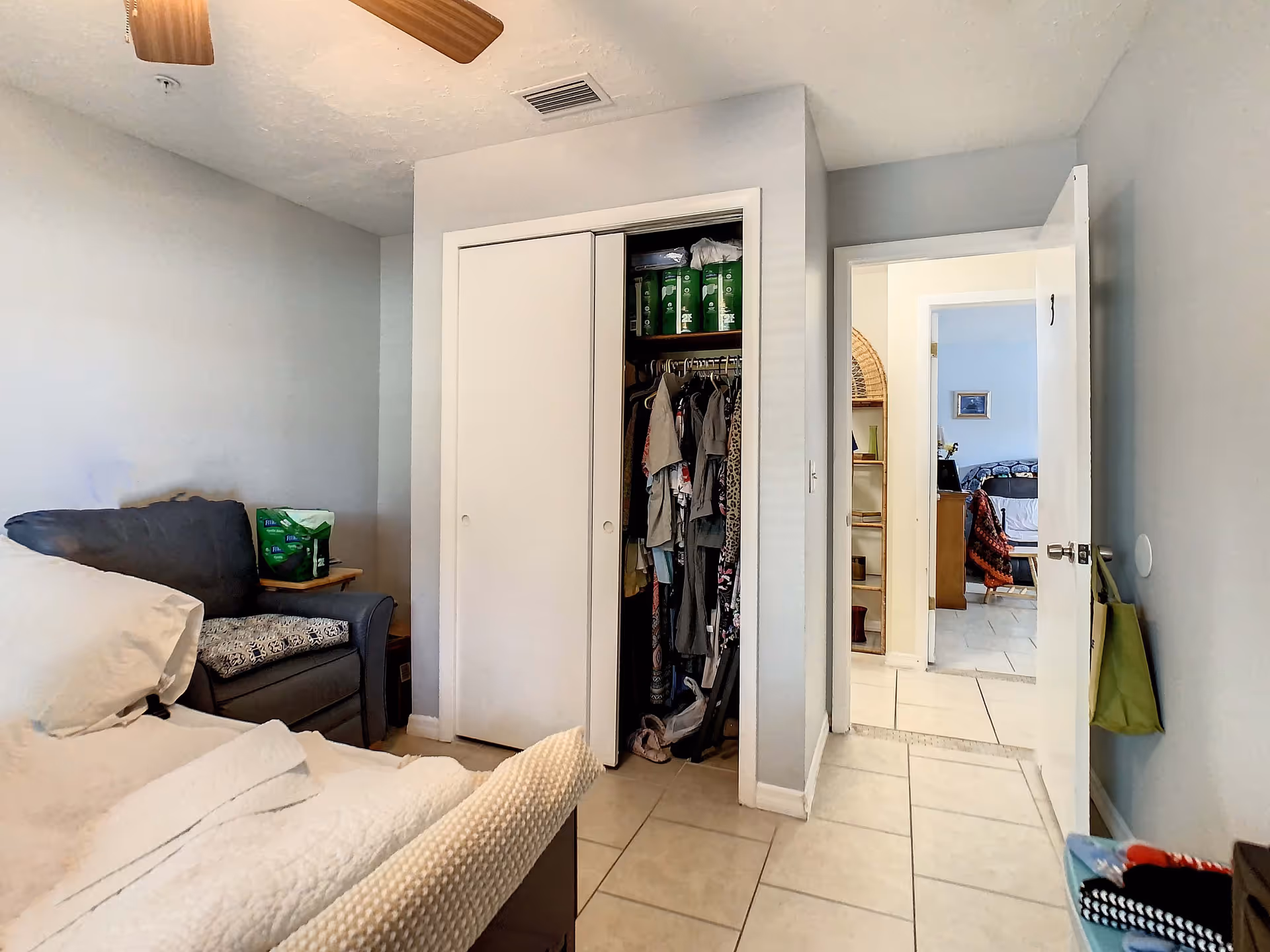A bedroom with a bed partially visible on the left, a gray armchair with a cushion and a pack of adult diapers on it, a closet with sliding doors partially open showing hanging clothes and items on the shelf, and an open door leading to another room with a wooden shelf and a bed visible in the background. The room has light-colored tiled flooring and light gray walls.