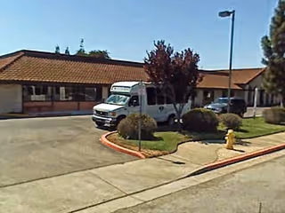 Exterior view of a single-story building with a tiled roof, a white van parked near the entrance, some bushes, a tree, a fire hydrant, and a streetlamp under a clear blue sky.