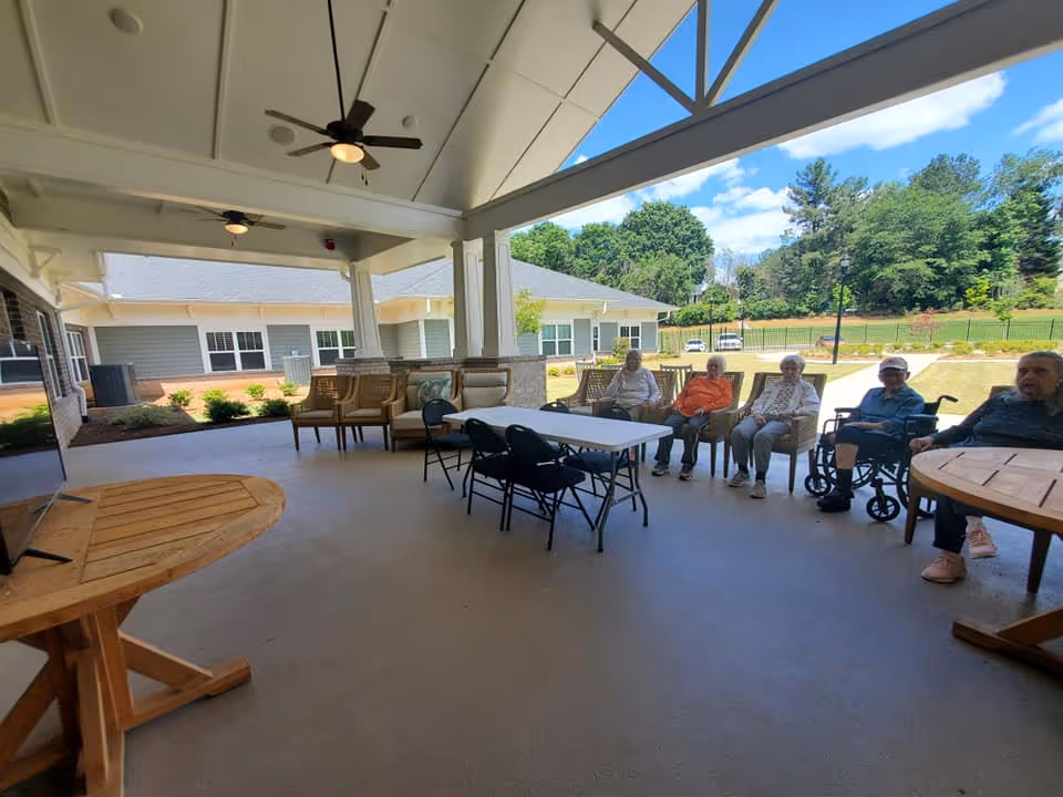Covered outdoor patio area with ceiling fans and seating, including chairs and tables. Five elderly individuals are seated along the edge of the patio, some in wheelchairs, with a view of a grassy area and trees in the background under a blue sky with clouds.