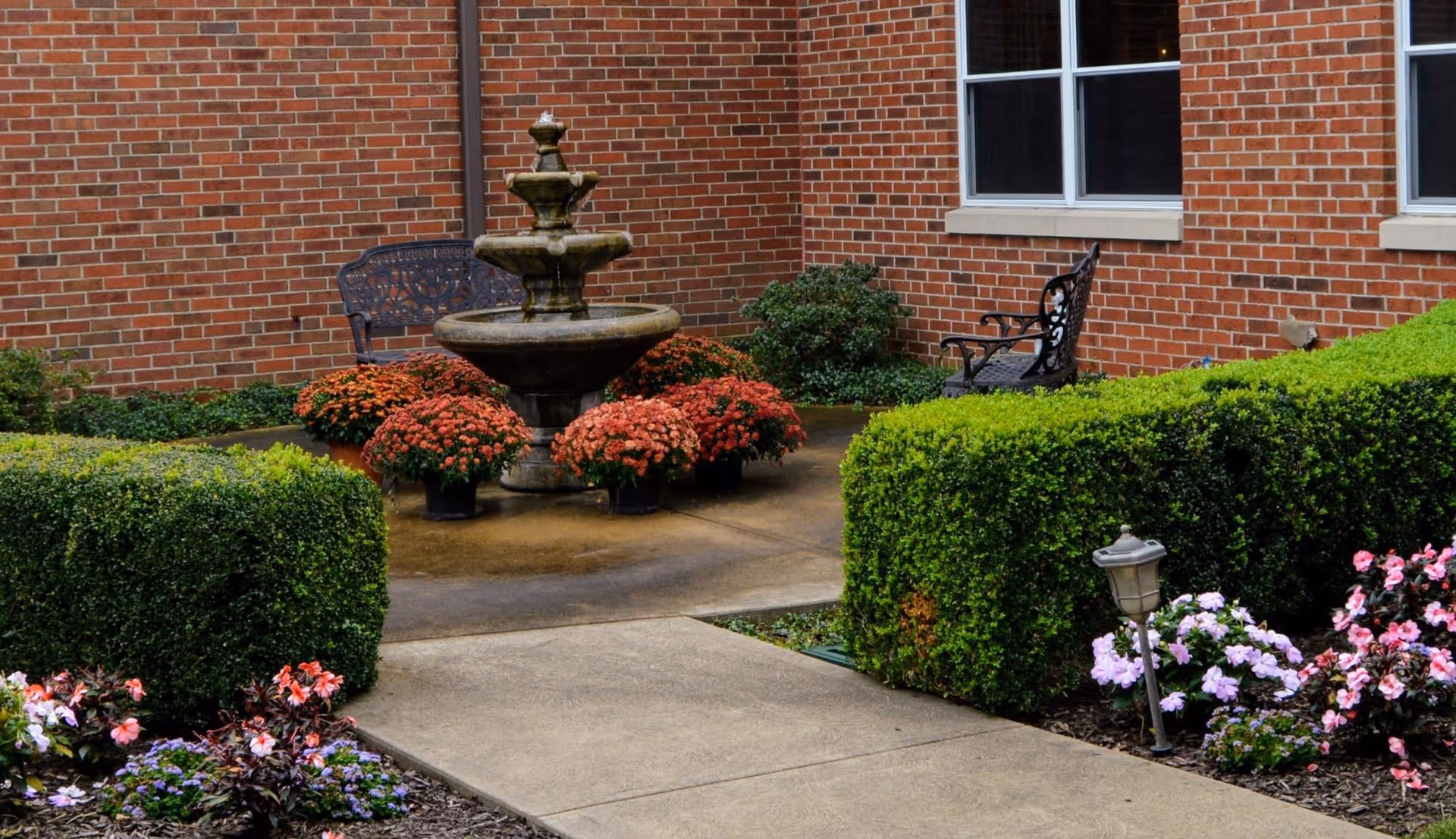 A small brick courtyard with a tiered fountain, benches, trimmed hedges, and flowering plants along a concrete pathway.