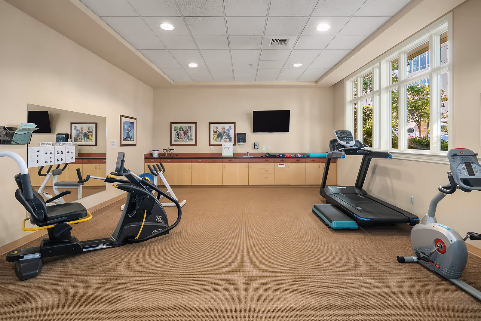 A fitness room with exercise equipment including a recumbent bike, treadmill, and stationary bike. The room has beige walls, a large mirror on one side, framed pictures on the wall, a flat screen TV mounted on the wall, and large windows letting in natural light.