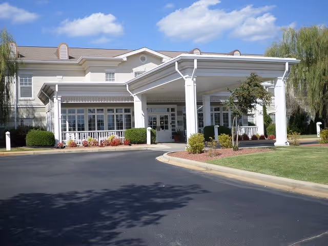 Front exterior view of a senior living facility with a covered entrance, white columns, large windows, and well-maintained landscaping including bushes and small trees under a partly cloudy blue sky.