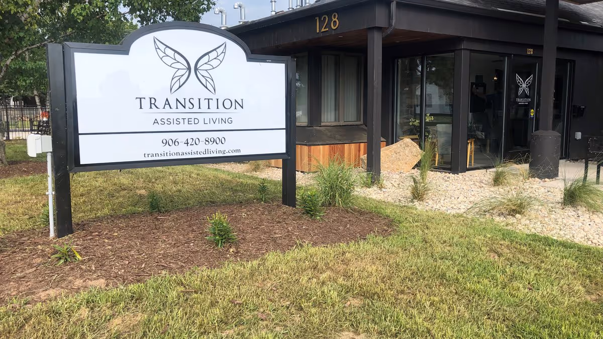 Outdoor view of Transition Assisted Living entrance with a large white sign showing the facility name, phone number and website in front of the building.