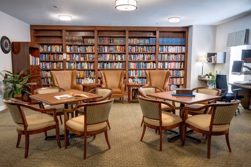 A cozy library room with two wooden tables surrounded by four cushioned chairs each. Behind the tables are three tan leather armchairs with small side tables between them. The back wall is lined with tall wooden bookshelves filled with books. To the right, there is a desk with a computer and office chair, a floor lamp, and some shelves with decorative items. A large plant is placed on the left side near a wooden cabinet.