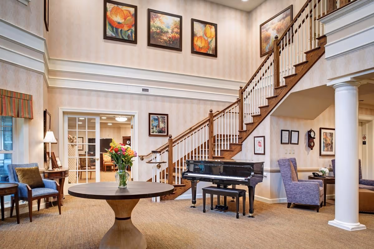 A spacious and elegant senior living facility common area featuring a grand piano, a round wooden table with a vase of colorful flowers, comfortable chairs, and a staircase with wooden handrails. The walls are decorated with framed floral and landscape paintings, and the room has warm lighting and beige carpeting.