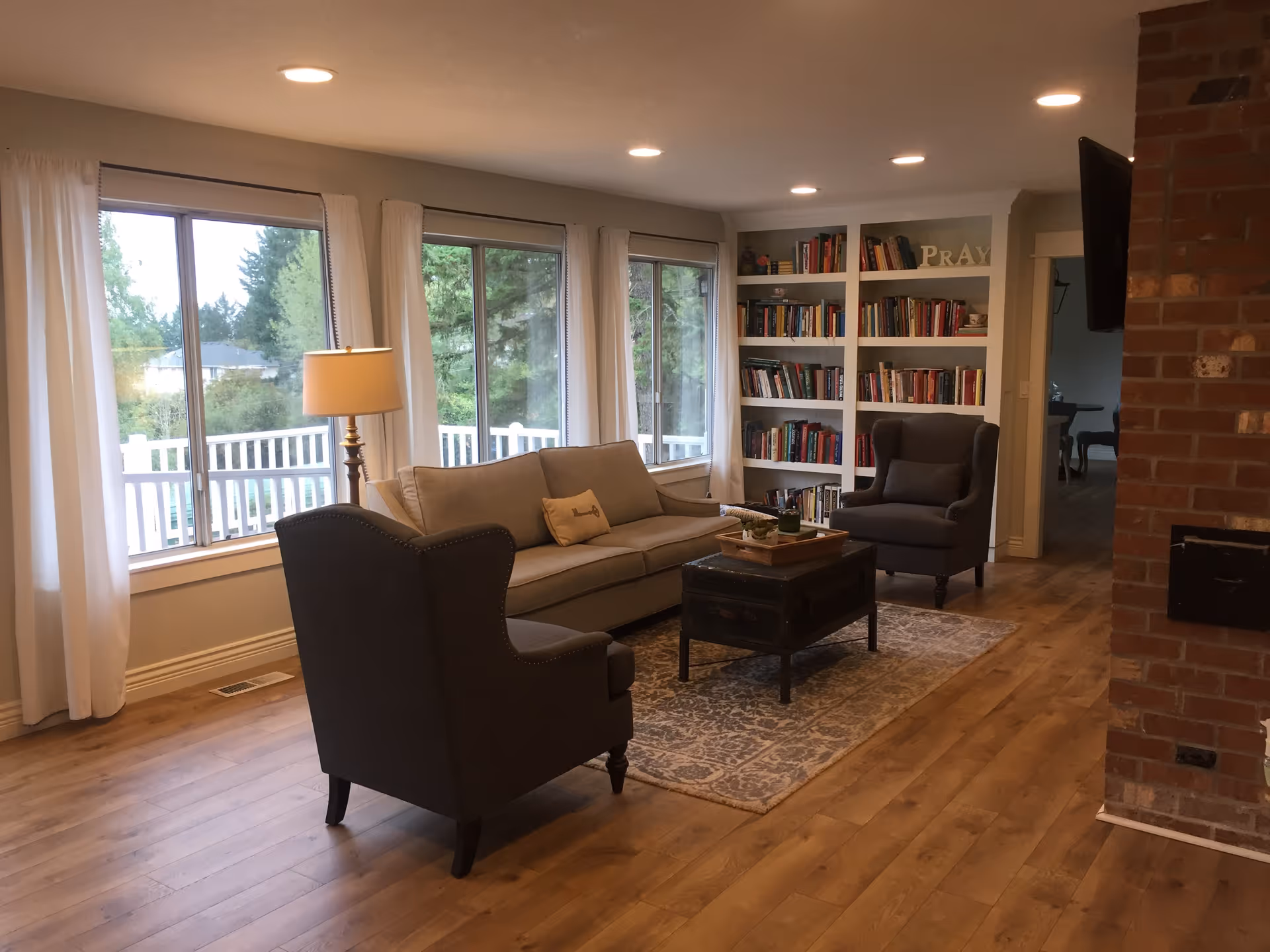A cozy living room with wooden flooring, a beige sofa, two dark armchairs, a wooden coffee table on a patterned rug, a floor lamp, large windows with white curtains showing green trees outside, built-in white bookshelves filled with books, and a brick fireplace on the right side.
