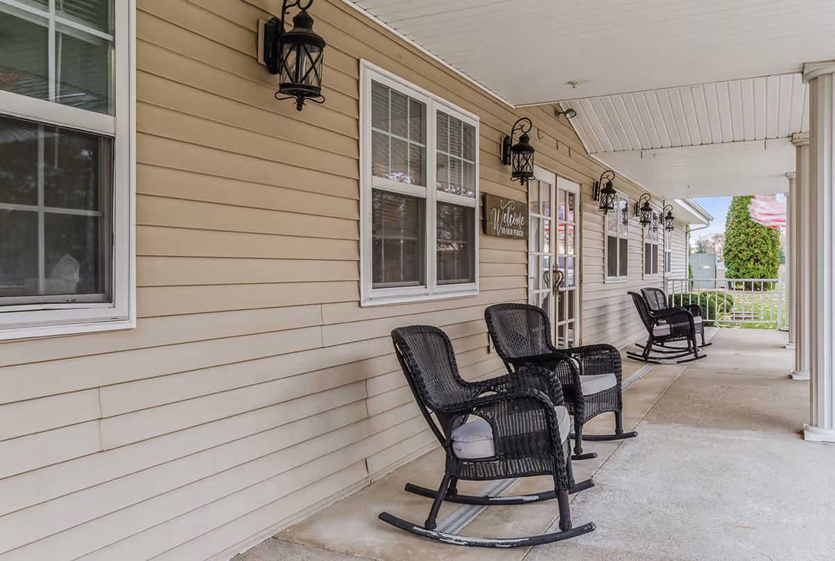 Covered porch area with beige siding, black wicker rocking chairs with cushions, multiple wall-mounted lantern-style lights, and a welcome sign near a glass door. The porch overlooks a grassy area with a tree and a flag in the background.