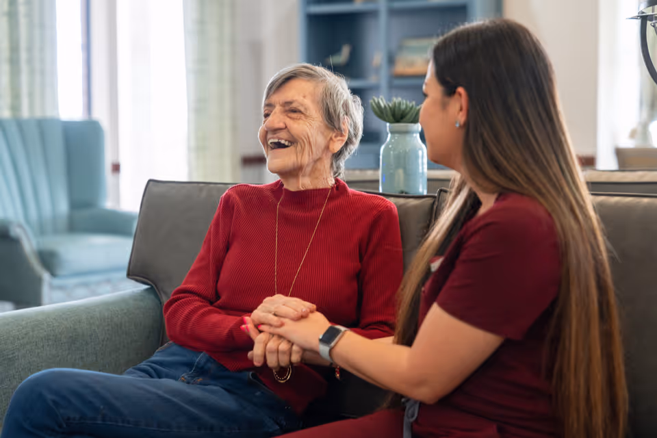 An elderly woman wearing a red sweater sits on a couch smiling and holding hands with a younger woman in a maroon uniform, who is sitting beside her in a cozy living room setting.
