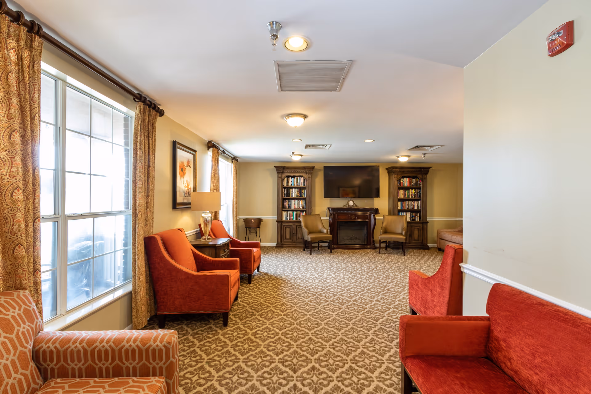 A spacious living room area with patterned carpet, beige walls, and multiple seating options including red and orange armchairs and a red sofa. Large windows with patterned curtains allow natural light to enter. At the far end, there are two wooden bookshelves flanking a fireplace with a mounted flat-screen TV above it. A table lamp and framed artwork are also visible.