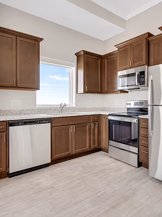 Bright modern kitchen with brown cabinets, stainless-steel appliances, a sink under a window, and light wood-look flooring.