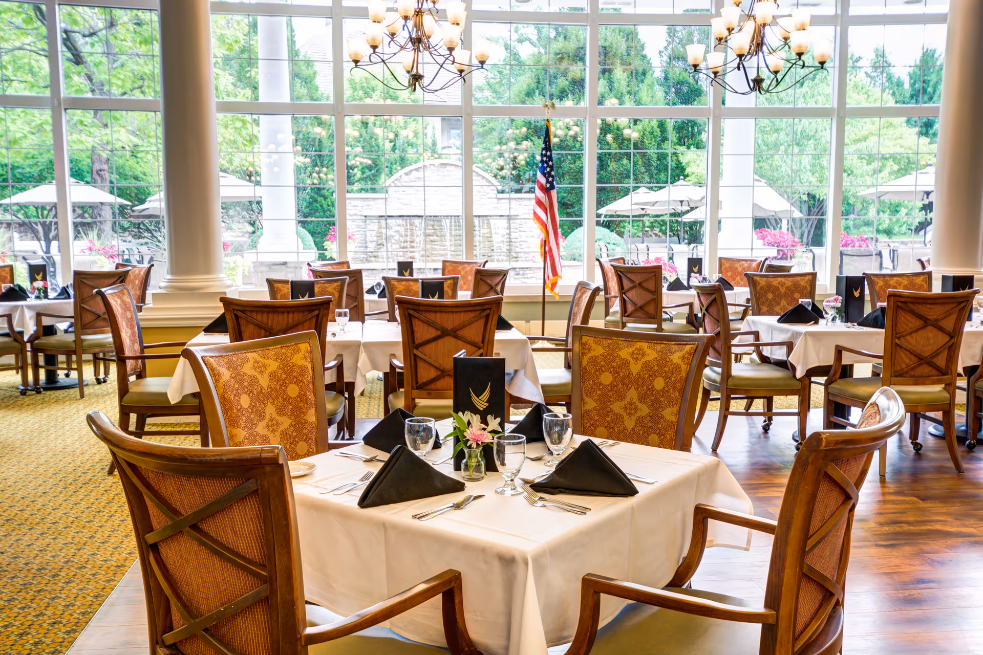 A bright and elegant dining room with multiple tables covered in white tablecloths, set with black folded napkins, glassware, and silverware. The room features large windows with a view of an outdoor patio area with umbrellas and greenery. There are chandeliers hanging from the ceiling and an American flag near the windows.