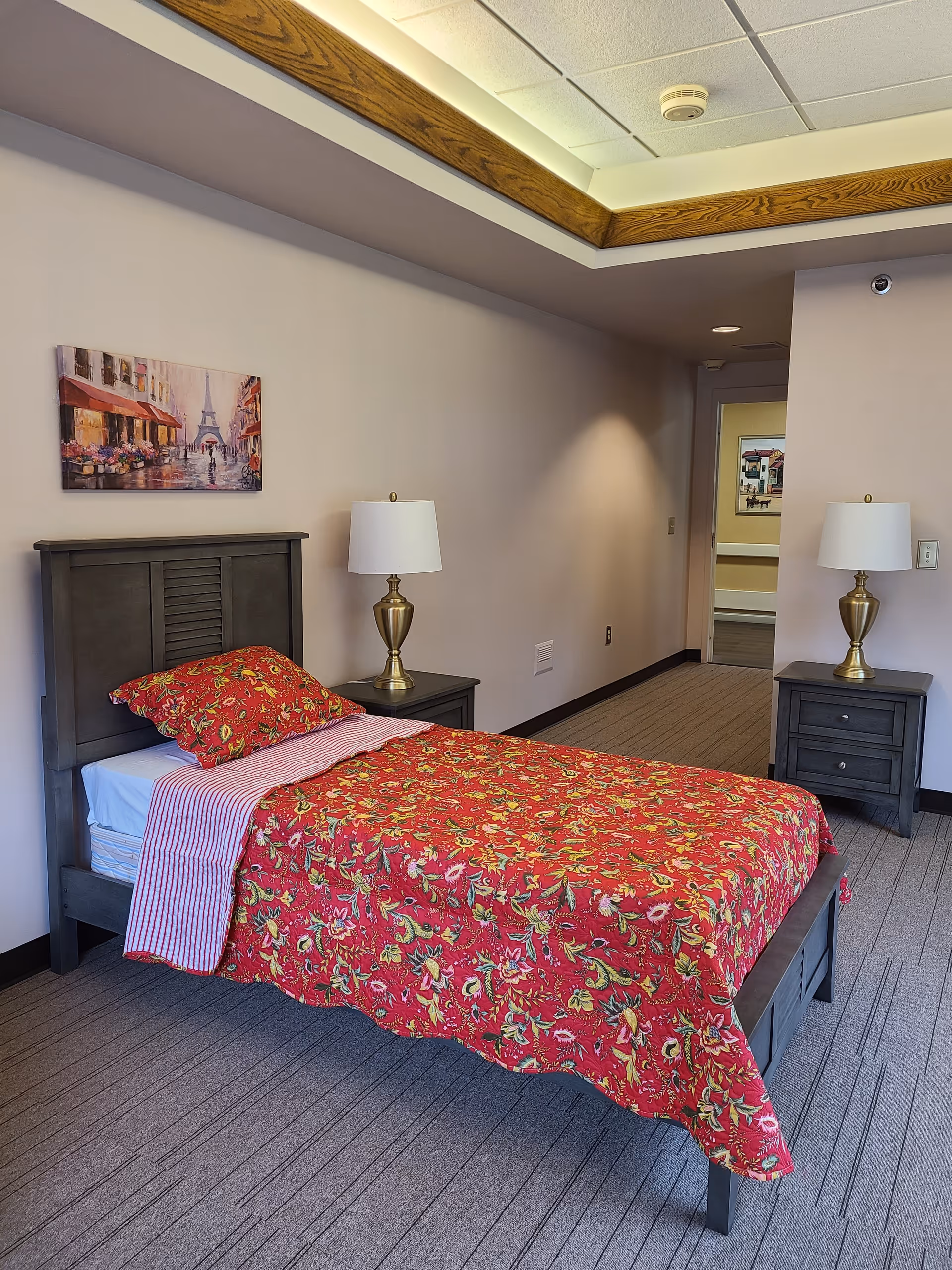 A bedroom with a single bed featuring a red floral bedspread and a striped sheet. The bed has a dark wooden headboard and is flanked by two matching dark wooden nightstands, each with a gold-colored lamp with a white lampshade. On the wall above the bed is a painting depicting a street scene with the Eiffel Tower in the background. The room has beige walls, a carpeted floor, and a ceiling with recessed lighting and wooden trim. A doorway leads to another room with a framed picture visible on the wall.