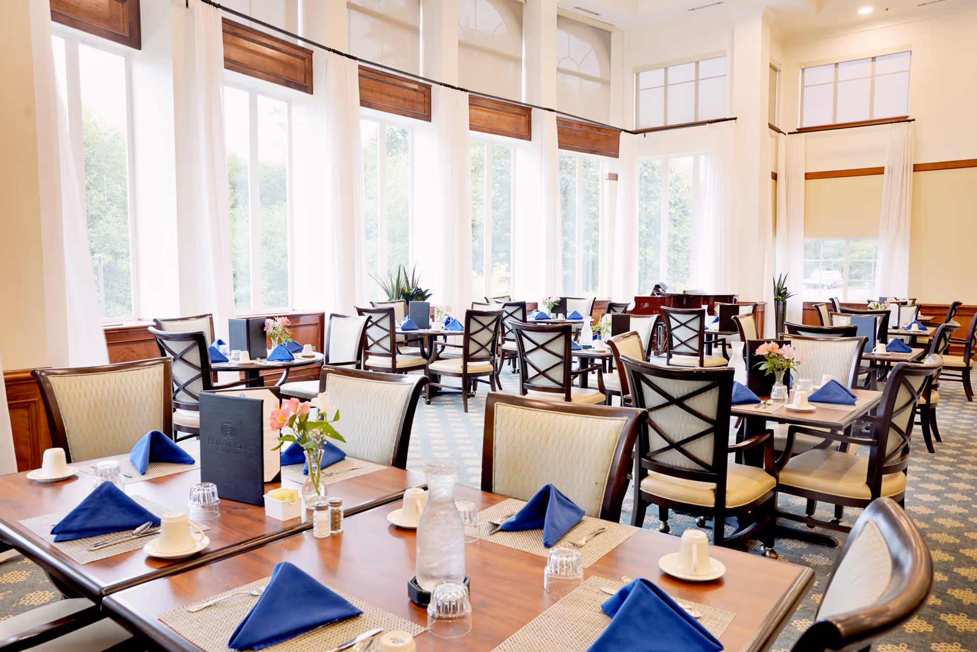 Bright and spacious dining room with large windows letting in natural light. Tables are set with blue napkins, white cups, glasses, and silverware. Chairs with cushioned seats and backs surround the tables. There are flower vases on the tables and a piano in the background.