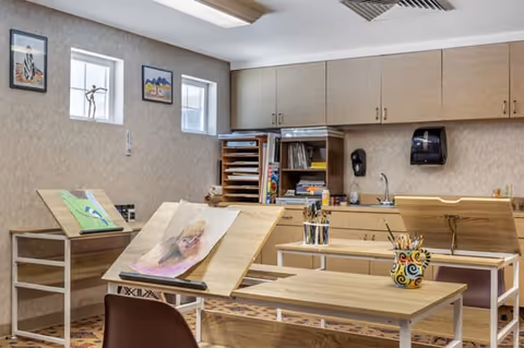 Well-lit art and craft room with adjustable drawing easels, tables, cabinets, and art supplies.