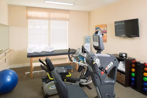 A small fitness room with exercise equipment including a recumbent bike and an elliptical machine. There is a blue exercise ball on the floor, a table with folded towels near a window with blinds, a wall-mounted TV, and a rack holding colorful dumbbells.