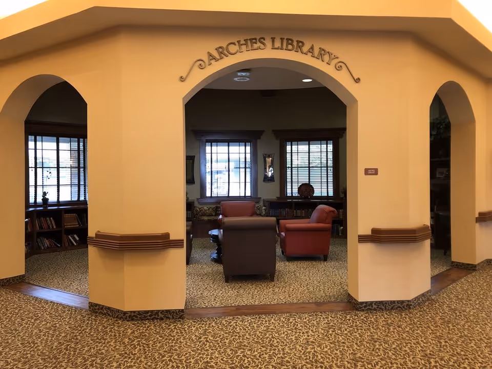 Interior view of a library room named 'Arches Library' with beige walls and carpeted floor. The room has three archways and contains comfortable seating including a red armchair, a brown armchair, and a sofa. Bookshelves with books are visible along the walls under windows with blinds.