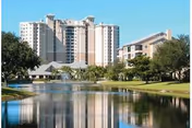View of a tall residential building complex named Arbor Trace, reflected in a calm body of water with trees and grassy areas surrounding the water under a clear blue sky.