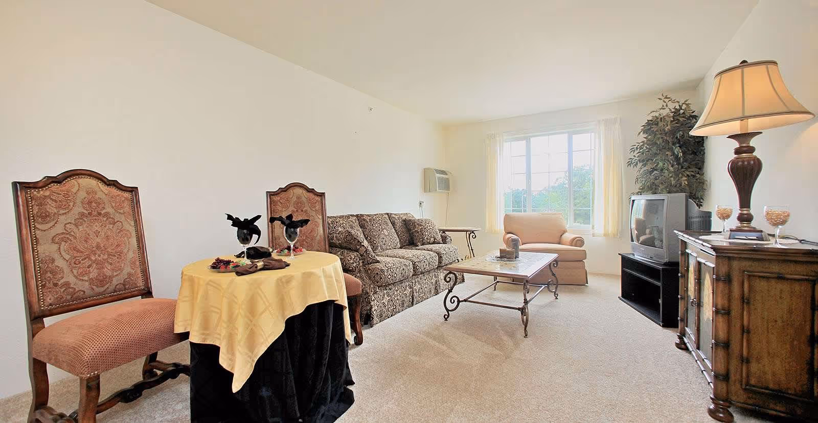 A bright living room with a large window letting in natural light. The room features a patterned sofa, a single armchair, a glass coffee table with a metal frame, and two ornate wooden chairs with a small round table covered by a yellow tablecloth. There is a wooden cabinet with a lamp and an older-style television on top, along with a tall plant in the corner.