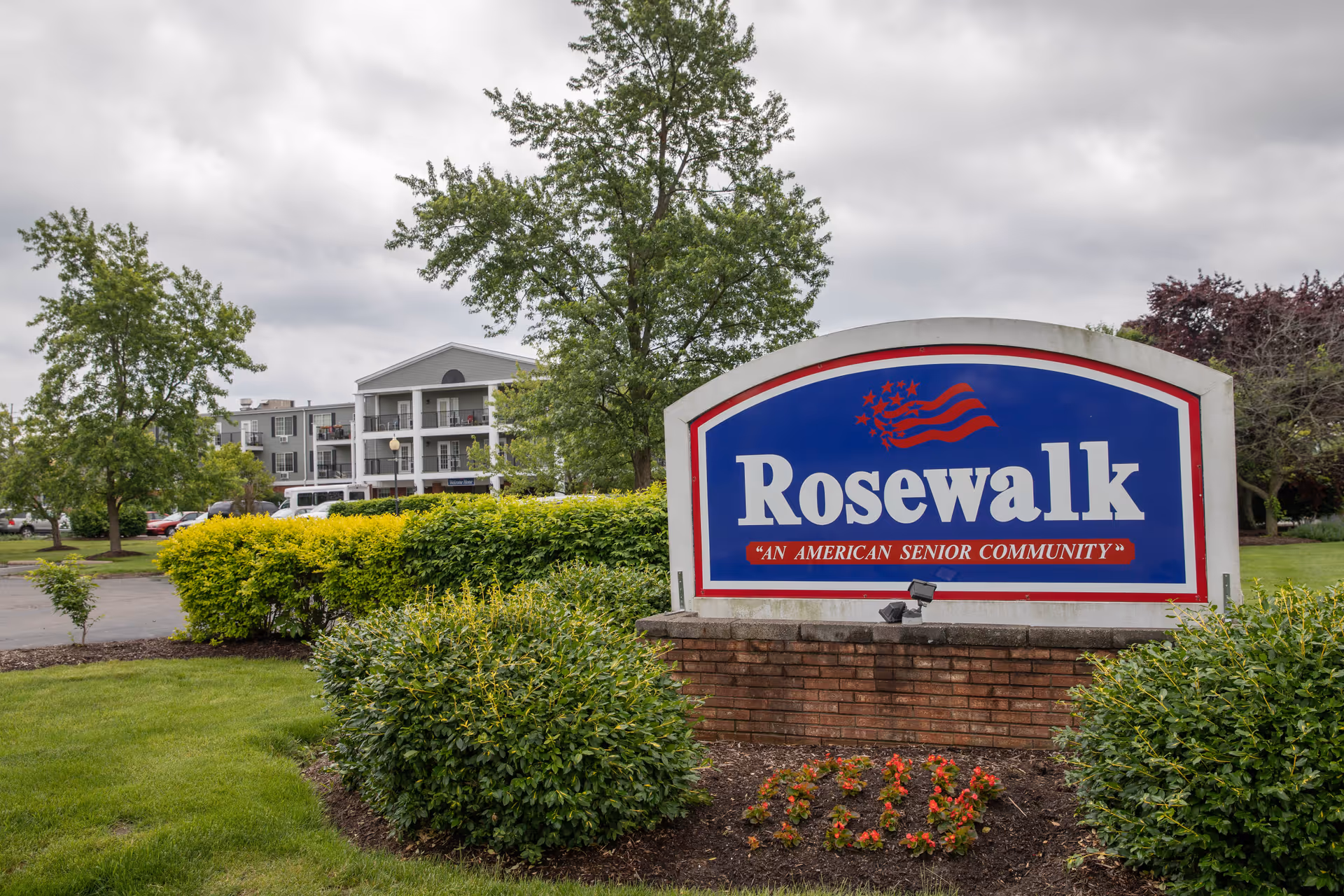Blue and red 'Rosewalk' entrance sign in landscaped grounds with bushes and the multi-story facility visible behind it under a cloudy sky.