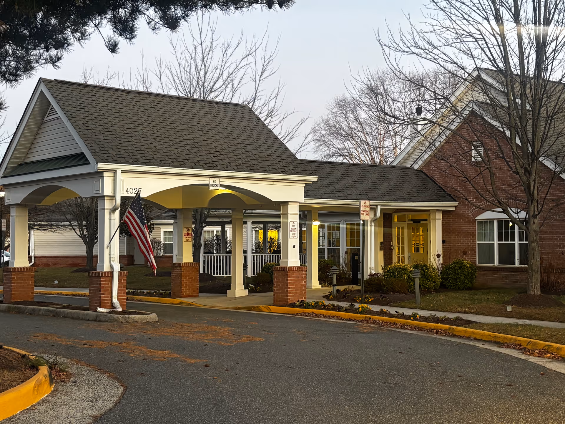 Entrance area of a senior living facility with a covered drop-off zone supported by white columns with brick bases. An American flag is hanging from one of the columns. The building has a brick exterior with windows and a door illuminated from inside. Leafless trees and some landscaping are visible around the entrance.