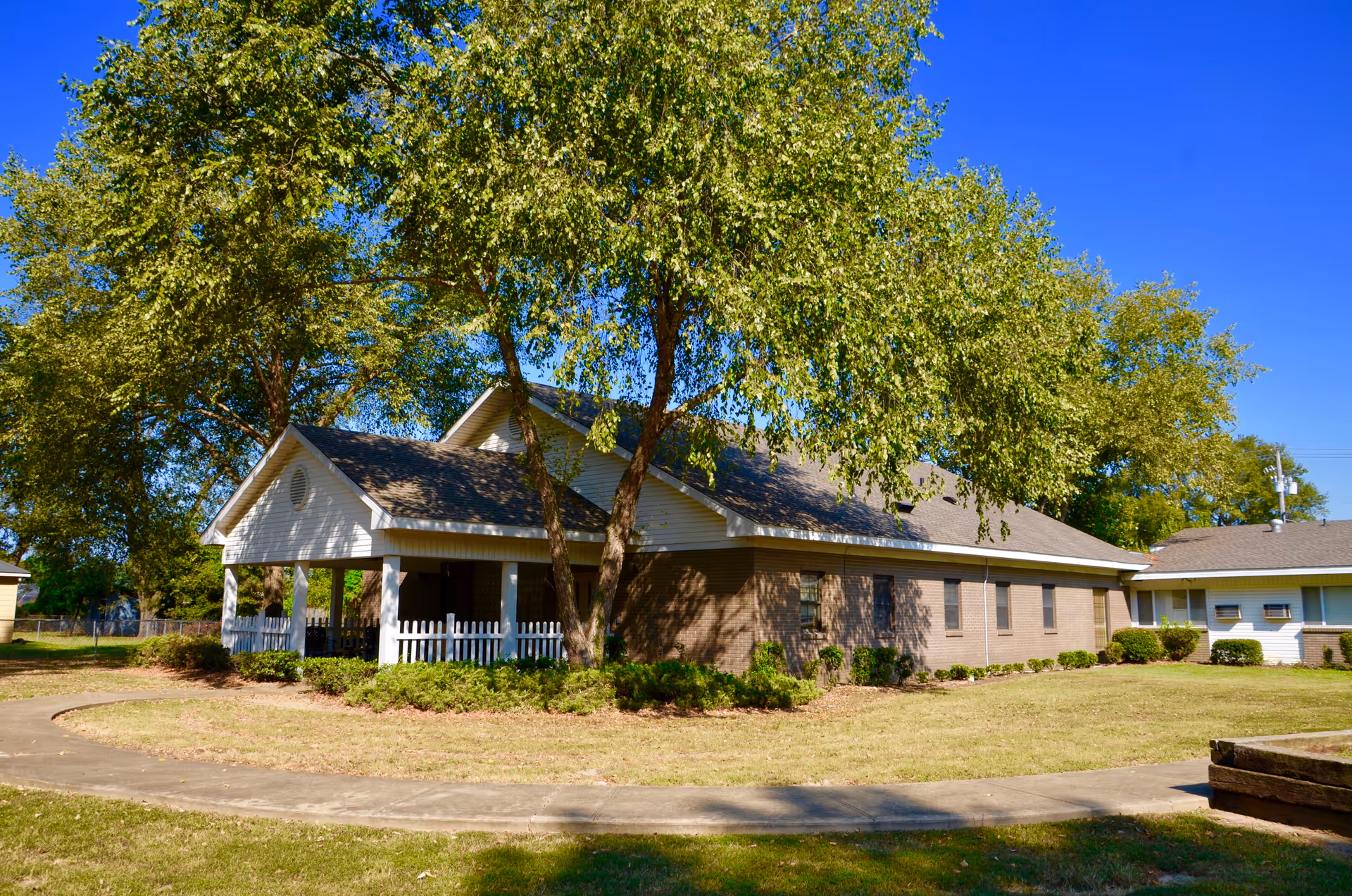 Single-story brick and siding building with a covered front porch, white picket fence, and large trees on a sunny lawn.