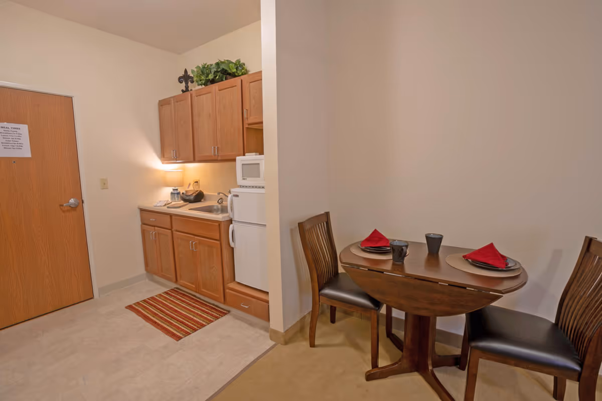 Small kitchenette area with wooden cabinets, a sink, a microwave, and a mini refrigerator. Next to the kitchenette is a small wooden dining table set for two with red napkins and black mugs. The floor transitions from tile in the kitchenette to carpet in the dining area.