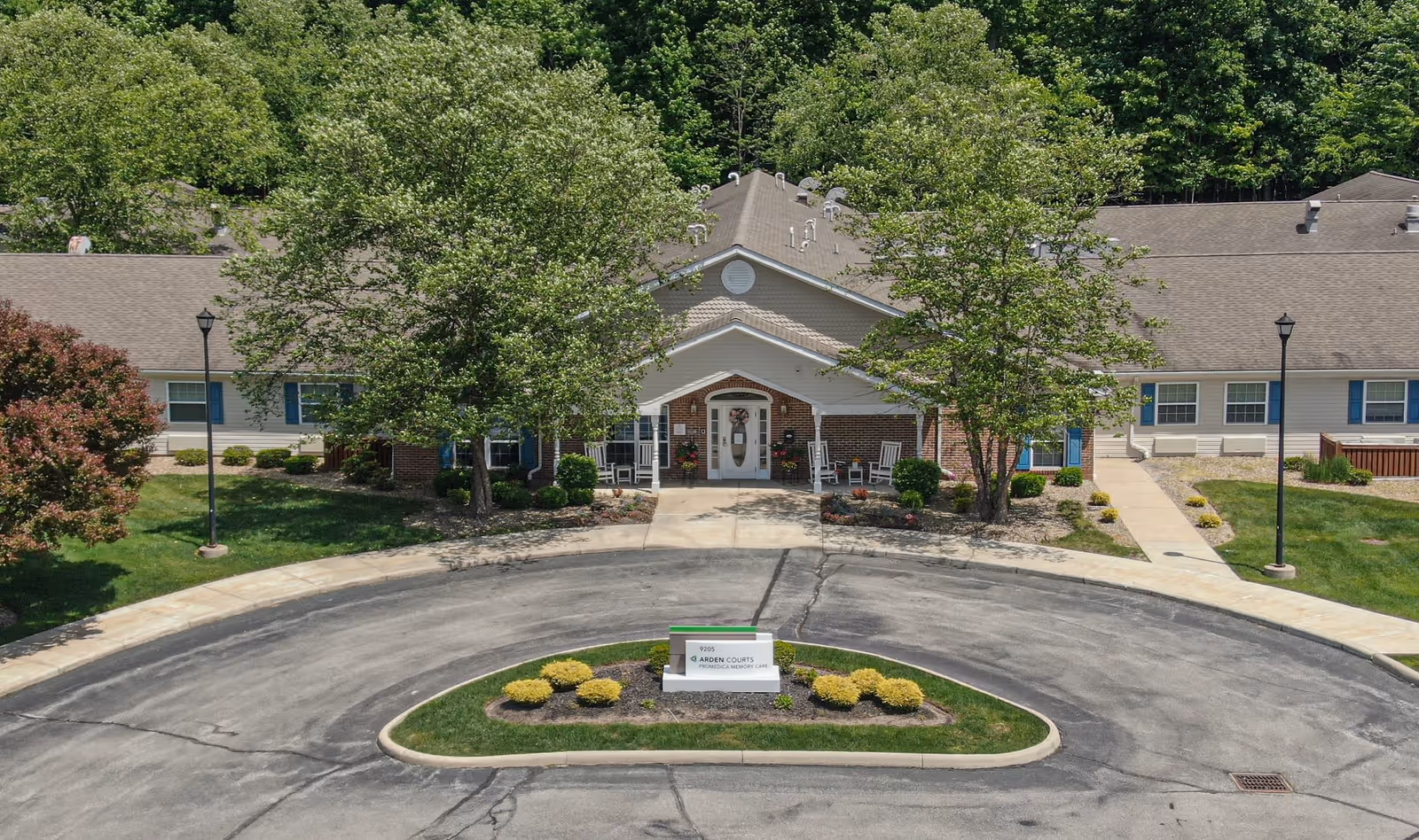 Front exterior view of Arden Courts - ProMedica Memory Care Community in Parma, showing a single-story building with a peaked roof, surrounded by trees and greenery. There is a circular driveway with a landscaped island in the center featuring a sign with the facility's name.