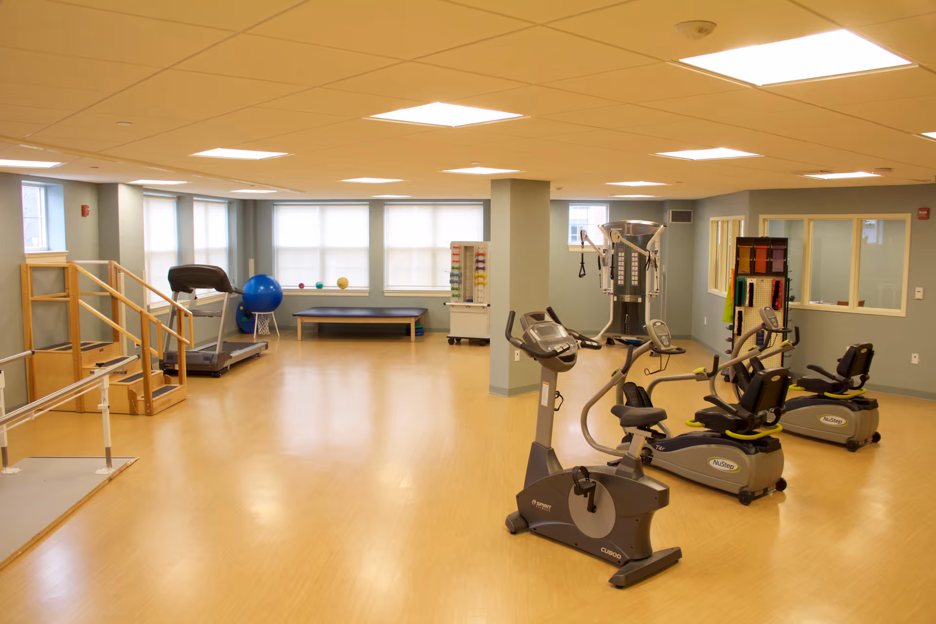 A spacious exercise room with light wood flooring and pale blue walls. The room contains various fitness equipment including stationary bikes, a treadmill, a multi-function exercise machine, a set of stairs with handrails, exercise balls, and a bench. Large windows with blinds allow natural light to enter the room.