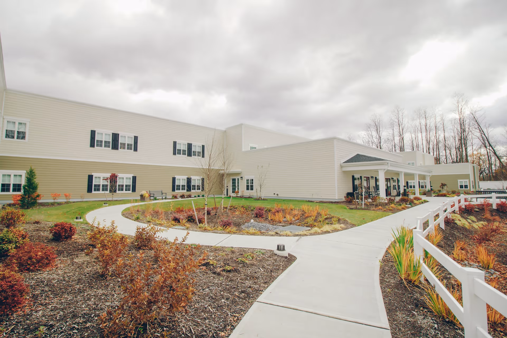 Exterior view of a senior living facility building with beige siding and black shutters, surrounded by landscaped gardens with shrubs and a curved concrete walkway leading to the entrance. The sky is cloudy and there are leafless trees in the background.