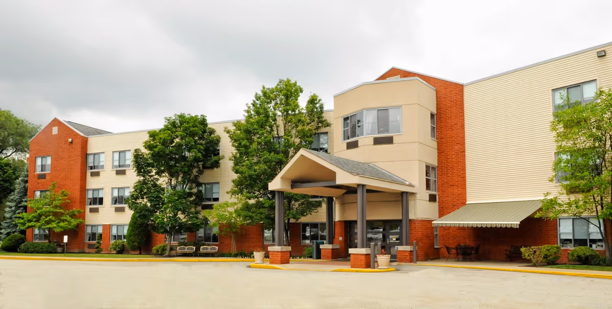 Exterior view of a three-story senior living facility building with a covered entrance, surrounded by trees and shrubs under a cloudy sky.