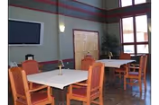 Community dining area with square tables and upholstered chairs, a wall-mounted TV, tall windows, and a cabinet.