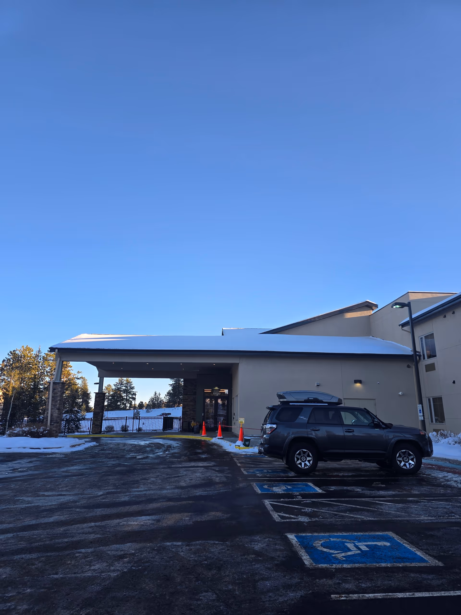 Exterior view of Forest Ridge Senior Living facility entrance with a covered drop-off area. A black SUV is parked in a handicapped parking spot in front of the building. Snow is visible on the ground and roof, with trees in the background under a clear blue sky.