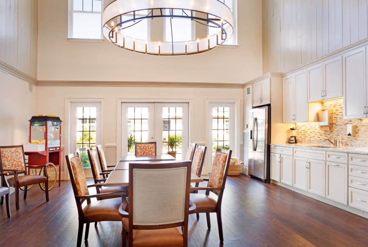 A bright and spacious dining area with a high ceiling and large windows. The room features a long wooden dining table surrounded by chairs with floral upholstery. To the right, there is a modern kitchen area with white cabinets, a stainless steel refrigerator, a coffee maker, and a sink. A popcorn machine is visible on the left side near the windows. The floor is dark wood, and a large circular chandelier hangs from the ceiling.