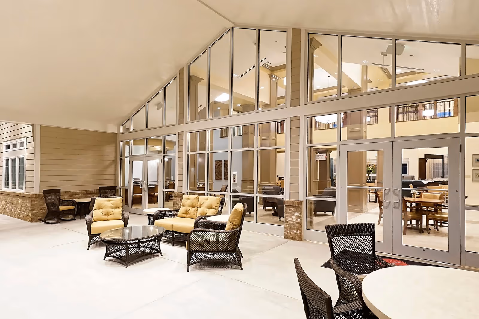 Outdoor seating area with cushioned wicker chairs and a glass-top coffee table on a covered patio. Large glass windows and doors reveal an interior space with tables and chairs, suggesting a common area inside the building.