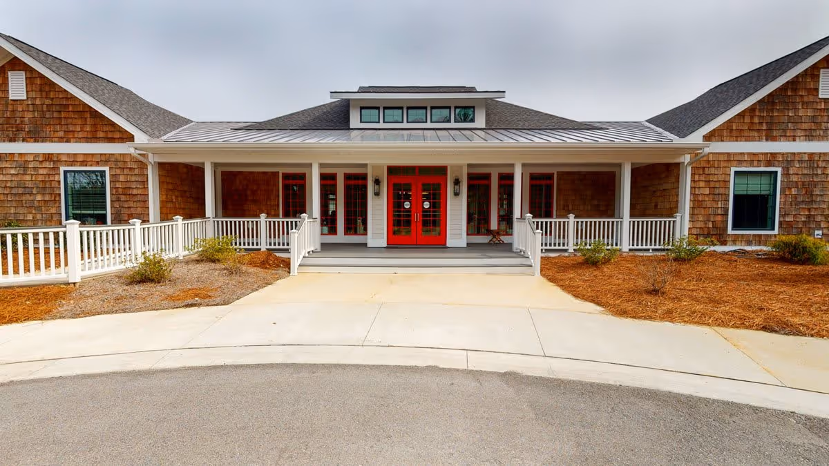 Front exterior view of a single-story building with brown shingle siding, white trim, and a metal roof. The entrance features bright red double doors with glass panels, flanked by windows and white railings on a small porch. There are landscaped areas with mulch and small shrubs on either side of the entrance driveway.