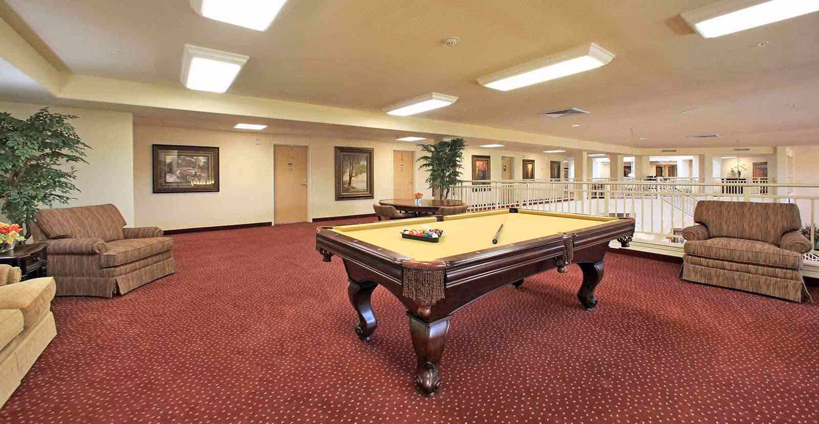A spacious indoor common area with a pool table in the center, surrounded by comfortable armchairs and small tables. The room has red carpet flooring, beige walls adorned with framed artwork, and ceiling lights. There are plants placed around the room and a white railing overlooking a lower level.