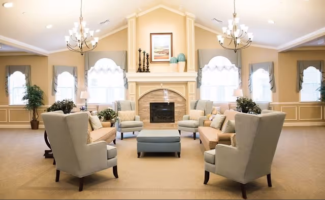 A spacious and well-lit living room with beige walls and carpet, featuring a central fireplace with decorative items on the mantel. The room has multiple windows with white curtains and valances, two chandeliers hanging from the ceiling, and comfortable seating including armchairs, sofas, and a central ottoman. There are also potted plants and table lamps adding to the cozy ambiance.