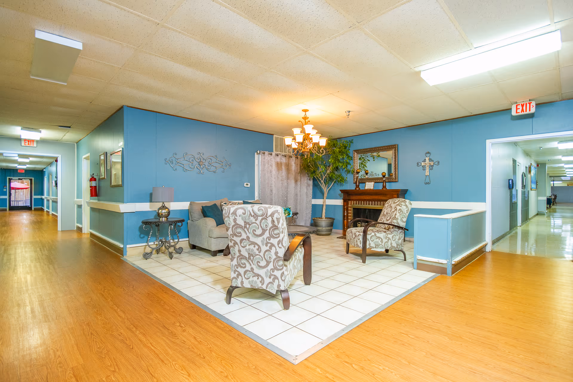 A cozy seating area in a senior living facility with two patterned armchairs, a beige sofa, a small table with a lamp, a decorative fireplace, a large mirror, a potted plant, and wall decorations. The seating area is tiled and surrounded by wood flooring, with blue walls and hallways extending on both sides.