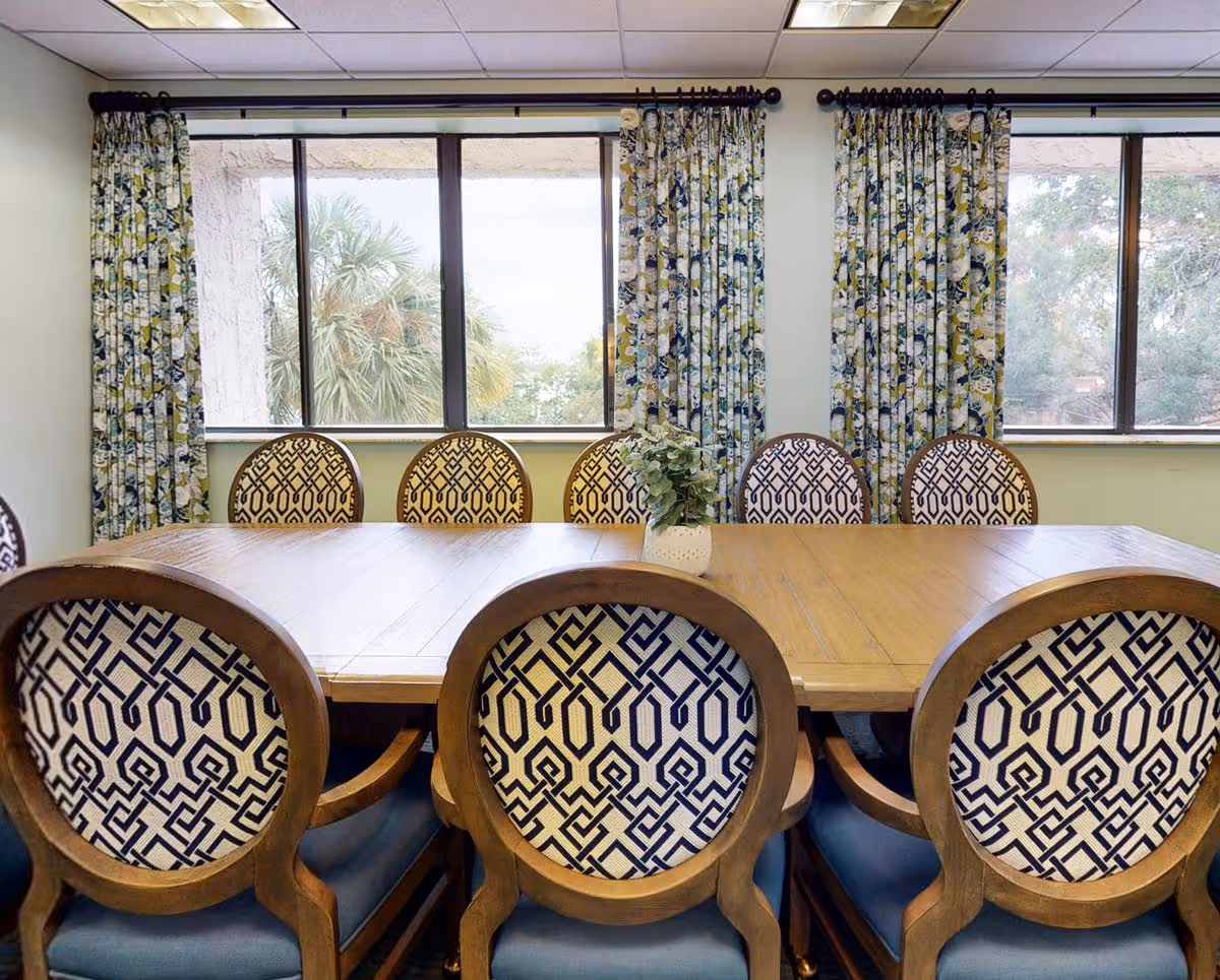 A dining room with a wooden table surrounded by eight chairs featuring patterned upholstery. Behind the table are three large windows with floral curtains, showing trees outside. A small potted plant is placed in the center of the table.