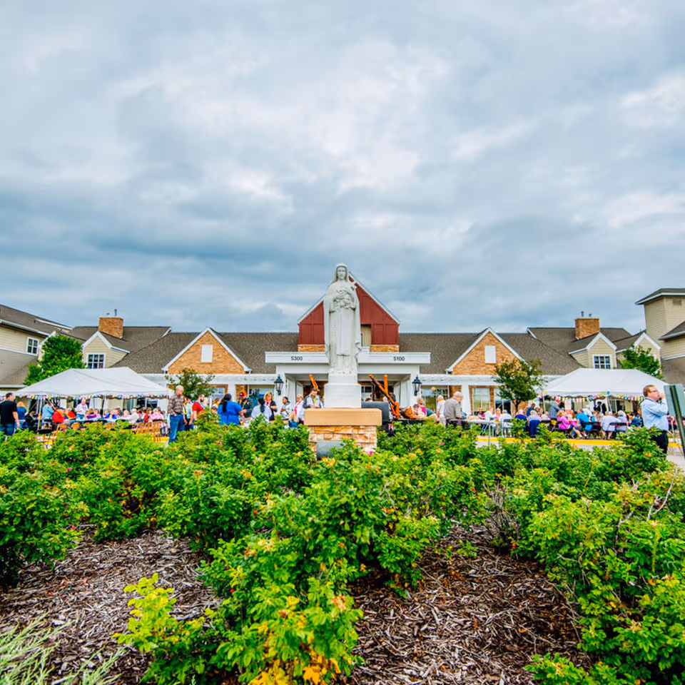 Outdoor view of Saint Therese Senior Living of Oxbow Lake featuring a statue in the center surrounded by green bushes and a crowd of people gathered under white tents in front of the building under a cloudy sky.